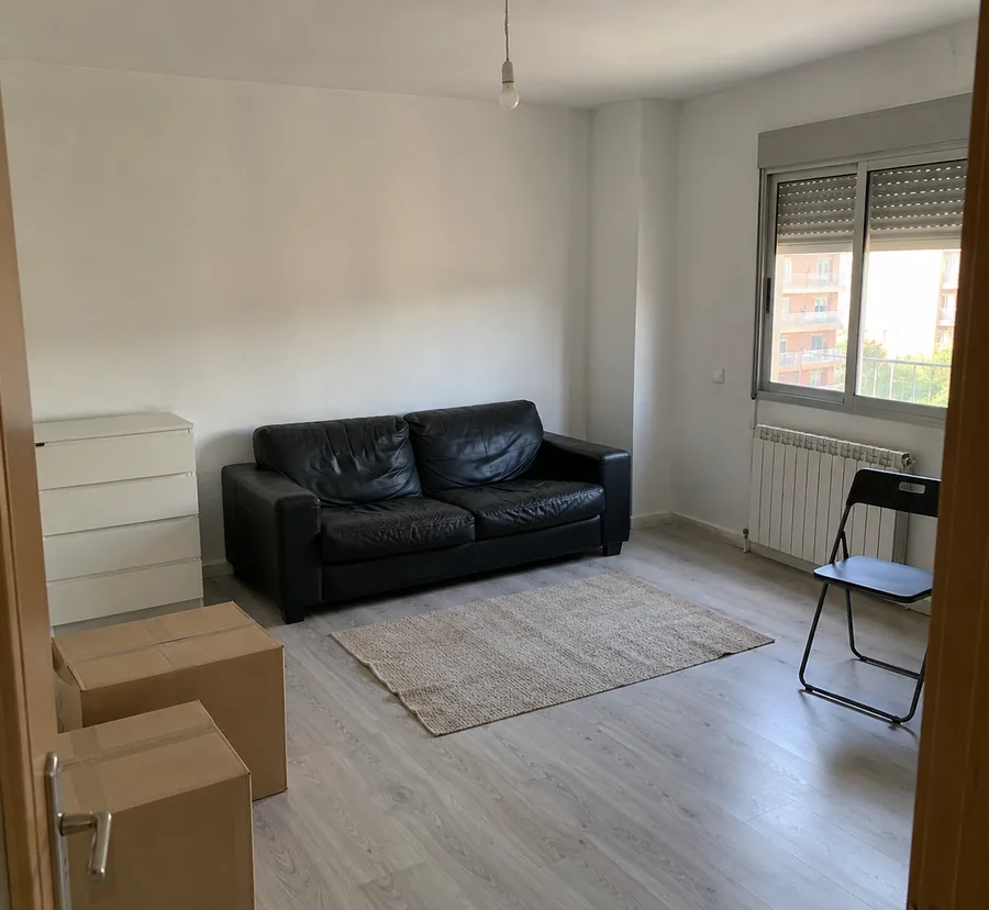 Salón a medio amueblar, foto hecha con el móvil desde la puerta: sofá negro viejo, cómoda blanca, cajas de mudanza, silla plegable y una bombilla sin lámpara